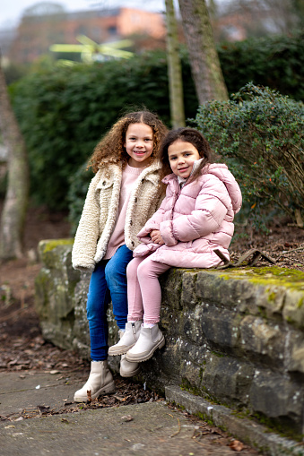 Full-length portrait of two young girls looking at the camera sitting on a wall in a public park. They are wearing warm casual clothing ready for a day out. The park is located in Gateshead.\n\nVideos are also available for this scenario.