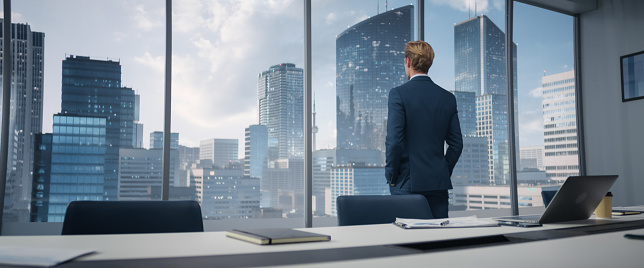 Caucasian Businessman Entering His Corner Office With Big City Megapolis View. Male Digital Entrepreneur Standing And Looking Out Of Window Thoughtfully, Planning Business Decisions.