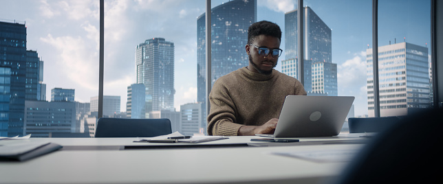 Confident Black Businessman Sitting at Desk in Modern Office, Using Laptop Computer, Next to Window with Big City with Skyscrapers View. Focused Finance Manager Planning Work Projects.