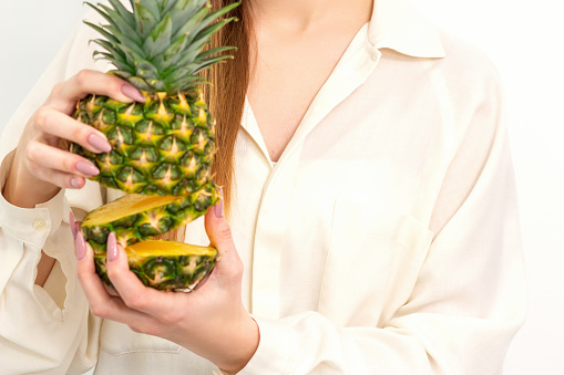 Beautiful young Caucasian woman holding pineapple and smiling, wearing a white shirt over white background.