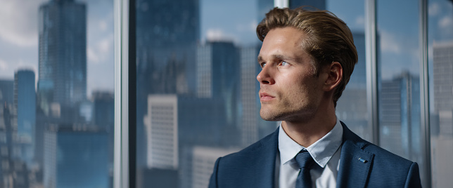 Portrait of Successful Caucasian Businessman in a Tailored Suit Standing in Office Looking out of the Window on Big City with Skyscrapers. Finance Manager Planning Project Strategy.