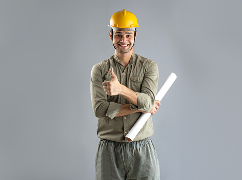 Portrait of smiling male architect holding blueprint showing thumbs up gesture while standing isolated on gray background