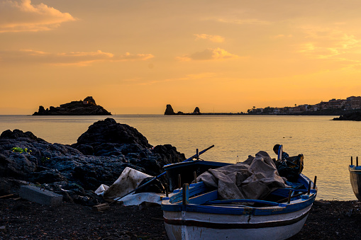 A traditional fishing beat is stranded on a pebble beach near Capo Mulini, in front of Isola Lachea, the famous Cyclops island in Homer's Odyssey, at sunset in Sicily, Italy