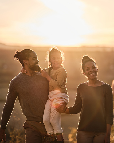 Happy African American parents and their small girl walking in nature at sunset. Copy space.