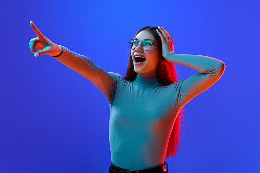 Wow. Surprised, delightful young girl, teenager in style glasses pointing that new sales season beginning isolated blue background in neon light. Concept of shopping, sales, fashion, style, emotions.