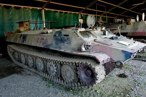 A close up on an old abandoned military truck, tank, or transporter made out of metal with rusty tracks attached to wheels seen on a dirt path located next to some logs, wood, spare parts, and forest