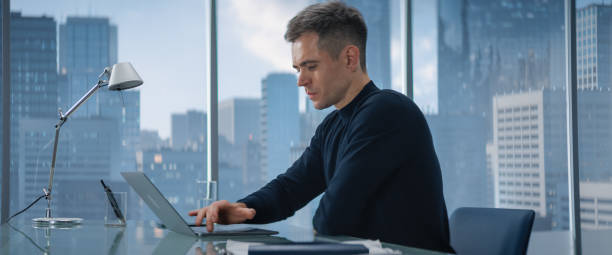 istock Creative Businessman in a Turtleneck Jumper Sitting in Modern Office, Using Laptop Computer Next to Window with Big City with Skyscrapers View. Finance Manager Planning Work Projects. 1798396507