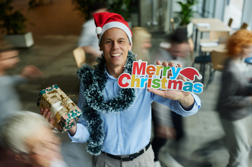 Happy mid adult businessman celebrating Christmas in the office and looking at camera. His colleagues are in blurred motion.