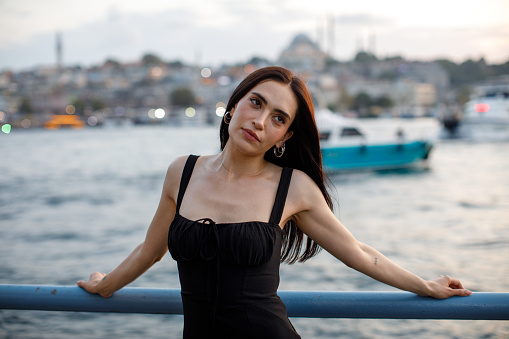 A stunning scene unfolds as a young and elegant woman, dressed in black, poses gracefully on Istanbul's iconic Galata Bridge against the backdrop of a vibrant sunset. Her dark hair and fair complexion beautifully complement the warm hues of the evening sky, creating a captivating moment of joy and serenity.
Woman, Beauty, Black Dress, Sunset, Istanbul, Galata Bridge, Elegant, Pose, Cityscape, Urban, Lifestyle, Joy, Serenity