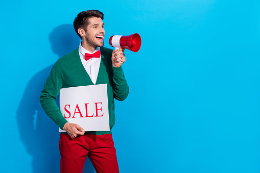 Photo of funny excited guy dressed xmas green cardigan holding sale card shouting toa empty space isolated blue color background.