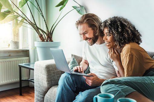 Multiracial young couple watching computer laptop sitting on the sofa at home - Happy diverse husband and wife using pc online services - Technology life style concept