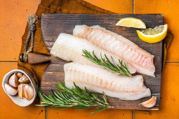 istock Raw cod fish fillets, codfish with rosemary on wooden board. Orange background. Top view 1798442482