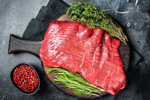Raw flank beef steak, uncooked meat with herbs. Black background. Top view