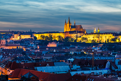 A view of Prague Castle at night lit up with golden lights, highlighting its Gothic architecture. The sky is a deep blue, and the trees in the foreground are dark and silhouetted against the skyline.