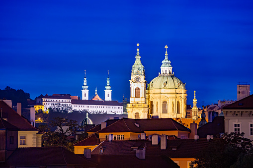 St. Nicholas Church in Prague lit up with white and yellow lights, creating a beautiful contrast against the deep blue sky. The buildings in the foreground are dark, with only a few lights visible.
