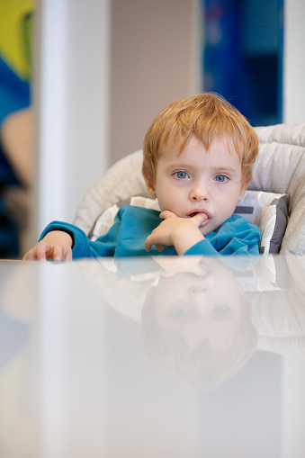 Blond toddler sitting in high chair and waiting for a meal, looking at camera with thumb in his mouth