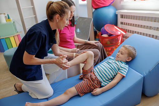 Female therapist working with preschool boy in the exam room, he is lying on his back and she is bending his legs in knee, holistic therapy for neurofunctional reorganization also known as Padovan Method