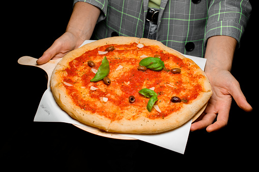 Waiter holds wooden board with fresh baked tasty Italian pizza with marinara sauce, garlic chips, olives, fresh basil leaves and dries herbs. Restaurant service, Waiter in gray uniform at work.
