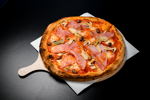 Italian pizza with bacon, mushrooms and olives on wooden serving board on black background. Top view. Italian restaurant menu photo.