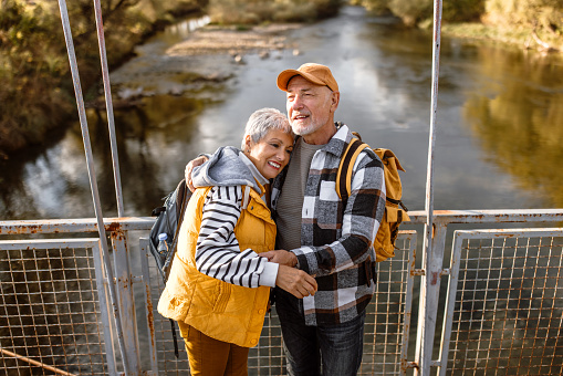 Active senior couple bonding in autumn nature walk, crossing the bridge over river.
