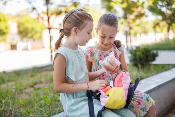 Two cheerful elementary age girls sitting outdoors on a school break, sharing sandwiches from backpack