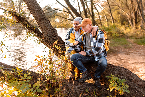 Active senior couple taking a break from walk, sitting next to the river, enjoying sunset and fresh autumn air.