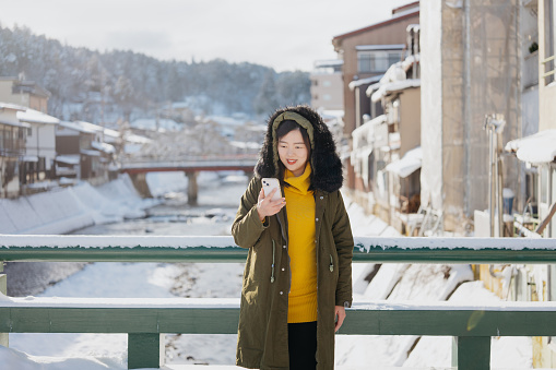 Asian pregnant woman standing in front of the bridge surrounding with snow and using mobile phone. Female traveller using smartphone during her winter holiday in Takayama, Japan.