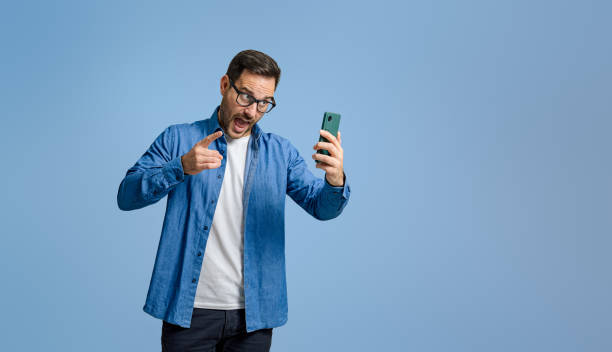 istock Businessman screaming angrily and pointing at smart phone while video calling over blue background 1798498340