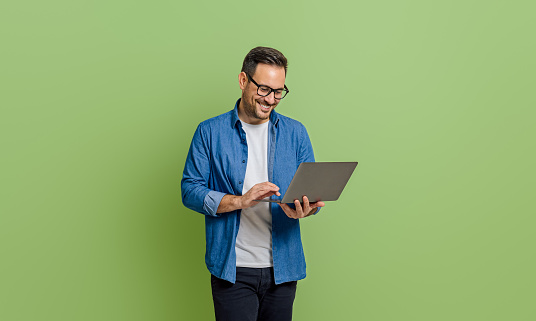 Smiling young businessman analyzing data over laptop while standing isolated on green background