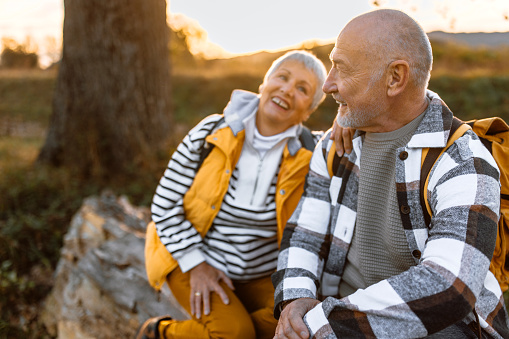 Active senior couple on a walk in a beautiful autumn nature, taking a break, sitting on a tree trunk, enjoying fresh air and beautifu view in sunset.