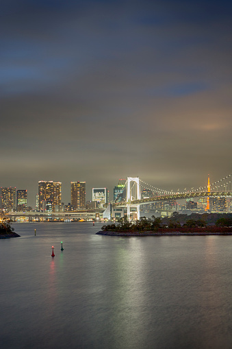 Asian Travel Destinations. Closeup View of Rainbow Bridge in Odaiba Island in Tokyo At Twilight with Line of Skyscrapers in Background. Vertical Shot