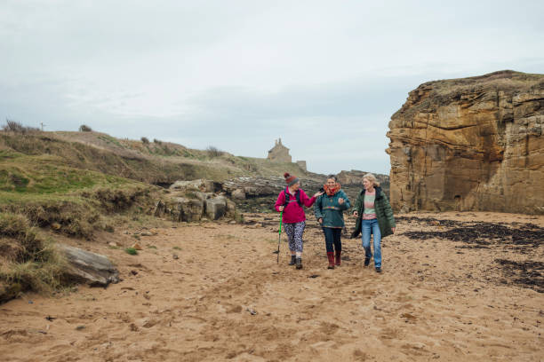 A wide angle view group of mature and senior women enjoying a day out in Bamburgh, North East England. They are enjoying a walk in the fresh air together while bonding. One of the older women is using a hiking pole on the multi terrain of the beach.