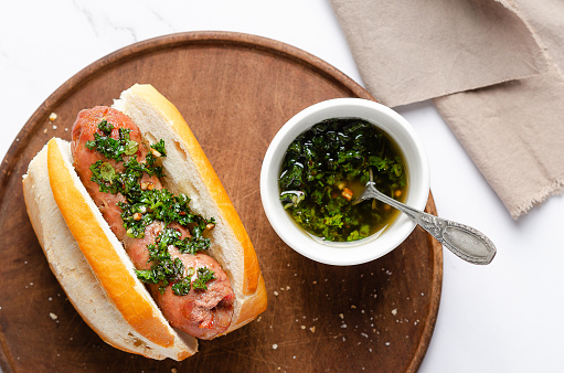 A choripan with chimichurri sauce on a wooden board and a brown napkin on white marble backgro