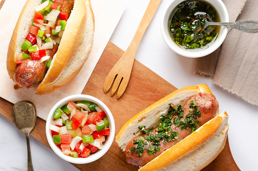 Choripanes with chimichurri and argentinian criolla sauce on a wooden board, chimichurri and criolla sauces in white bowls, two spoons, a fork and a brown napkin on white marble background.