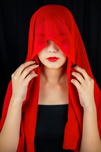 Portrait of a serene young woman covering her head with red dupatta and looking down with a blank expression over dark black background.