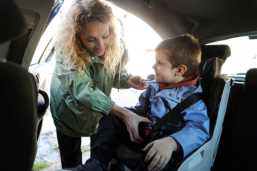 Boy sitting in a car seat for kids with his mother adjusting the belt. About 5 years old male and 30 years old woman, both Caucasian.