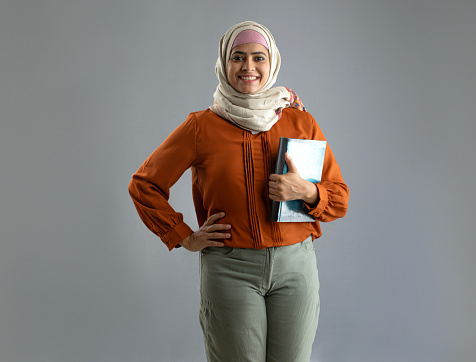 Portrait of smiling female student dressed in hijab holding book while standing over gray background