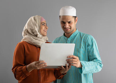 Portrait of cheerful young couple dressed in hijab and skull cap working on laptop standing against gray background