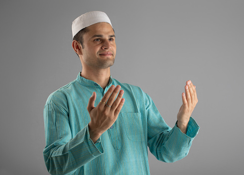 Portrait of man dressed in skull cap with hands raised praying and looking away while standing over gray background