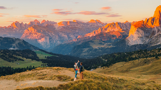 Drone high-angle  of woman and man with their dog giving High Five on the top of the mountain admiring the view of beautiful valley with hairpin curve road at Sella pass and the distant view of Mt. Marmolata - the largest mountain of Italian Dolomites - UNESCO World Heritage Site, during colourful heaven-like sunset.
