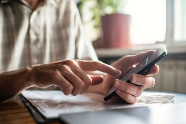 Senior man with smartphone, laptop and bills, documents or receipts at home