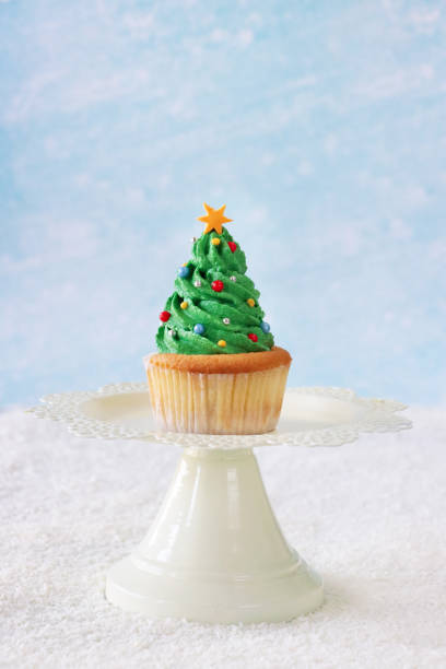 Stock photo showing close-up view of a cake stand displaying an individual, homemade Christmas tree design cupcake, in paper cake case, on icing sugar snow against a pale blue background. The cake is topped with swirl of green butter icing and decorated with multicoloured sugar sprinkles. Home baking concept. imagen de magdalena individual, casera, de diseño de árbol de navidad en soporte de pasteles, remolino de crema de mantequilla verde decorado con chispas de azúcar multicolores, nieve de azúcar glas, fondo azul pálido, enfoque en primer plano