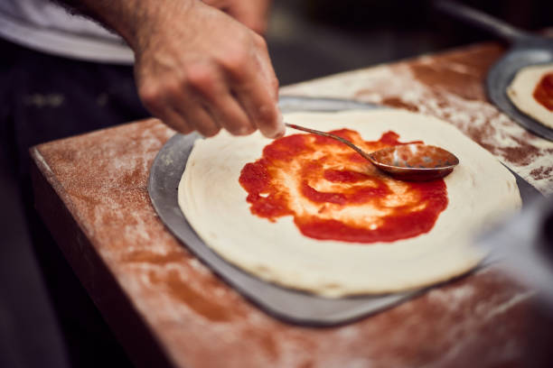 A man using a spoon, spreads tomato sauce over the pizza dough.