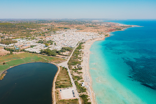 Drone high-angle photo of crystal blue yellow sand beach with groups of people and colourful umbrellas on the sand spit and the water reservation in South Puglia, Lecce Province, Ionian Italy