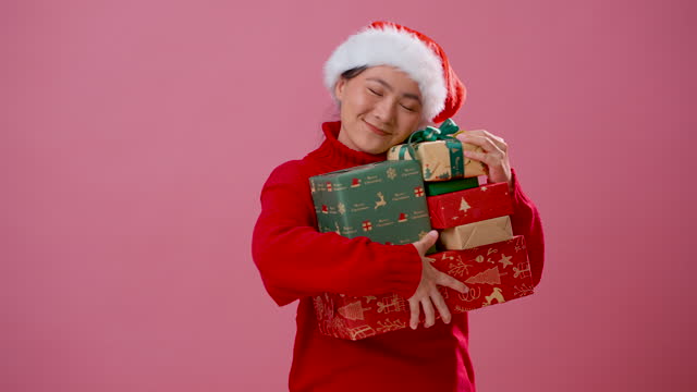 Asian woman wearing Santa hat holding many gift boxes standing isolated over pink background.