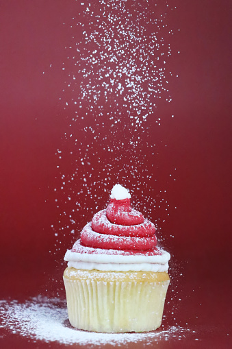 Stock photo showing close-up view of a freshly baked, homemade Father Christmas hat design cupcake, in paper cake case, displayed surrounded by icing sugar snow on red background. The cake is topped with a swirl of white and red butter icing. Home baking concept.
