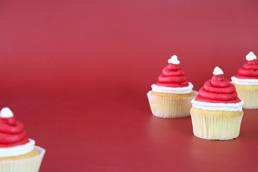Stock photo showing close-up view of a batch of freshly baked, homemade Father Christmas hat design cupcakes, in paper cake cases, displayed on a red background. The cakes are topped with swirls of white and red butter icing. Home baking concept.