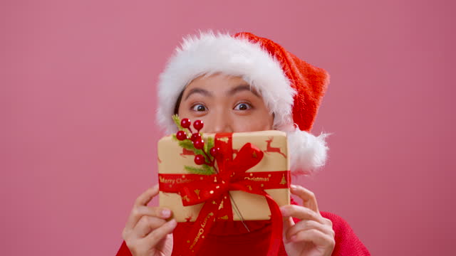 Asian woman wearing Santa hat showing a gift box covering her face, standing isolated over pink background.