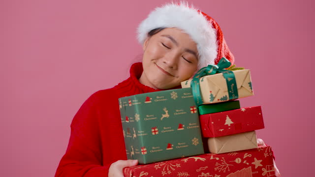 Asian woman wearing Santa hat holding many gift boxes standing isolated over pink background.