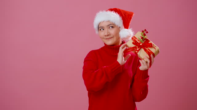 Asian woman wearing Santa hat happy smiling and shaking a gift box standing isolated over pink background.